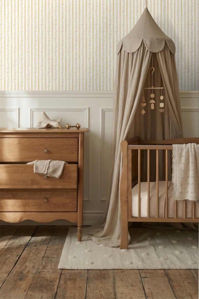 Vliestapete mit gestreiftem Blumenmuster in einem Babyzimmer mit Holzkommode, Gitterbett und Baldachin.