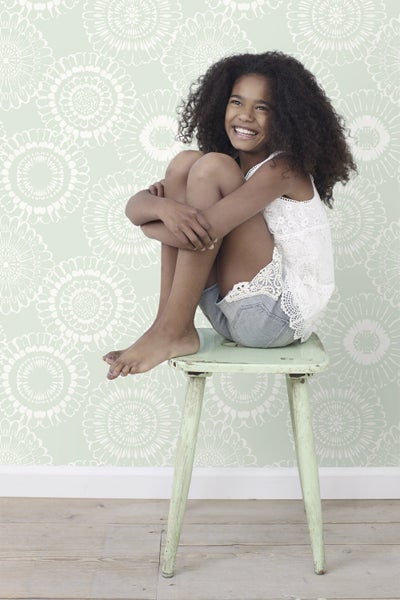 Lachendes Mädchen mit lockigem Haar sitzt auf einem Hocker vor einer Wand mit Blumenmuster