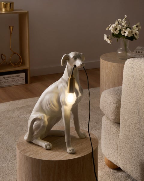 Weiße Tischleuchte in Form eines sitzenden Hundes, der ein Kabel mit goldener Fassung und klarem Leuchtmittel im Maul hält.