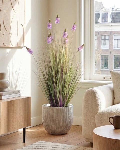 Künstliches Ziergras mit lila Blüten in einem hohen, strukturierten Topf, platziert in einem hellen Wohnzimmer neben einem Fenster.