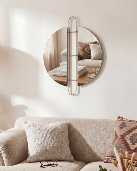 Runder Wandspiegel mit vertikalem Metallrahmen, der zwei Glashalbkreise trennt, hängend über einem Sofa in einem hellen Wohnzimmer.