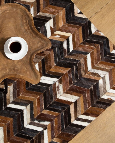 Patchwork-Teppich aus Kuhfell in Braun, Schwarz und Weiß mit Chevron-Muster neben einem rustikalen Holztisch mit Kaffeetasse.