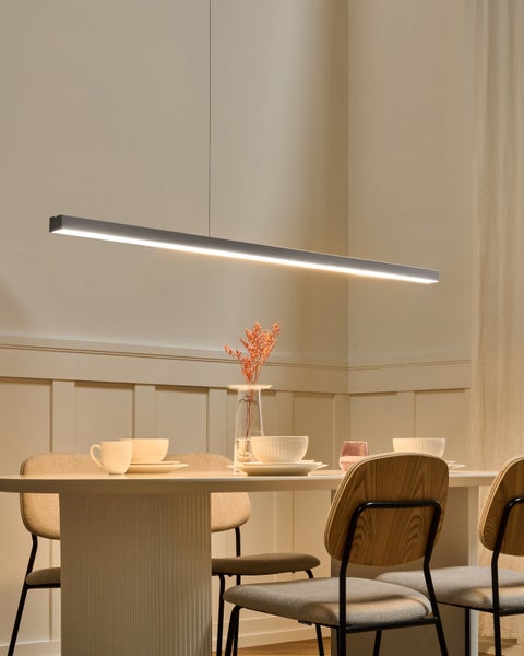 Moderne lineare LED-Pendelleuchte über einem gedeckten hellen Esstisch mit Holzstühlen in einem hellen minimalistischen Raum.