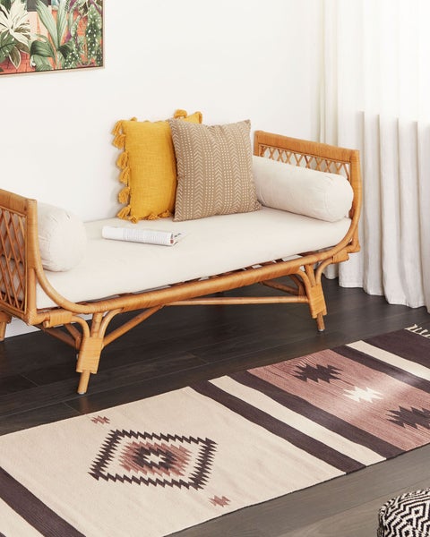 Daybed aus Rattan mit hellen Polstern und Zierkissen in einem hellen Raum mit dunklem Holzboden und gemustertem Teppich.