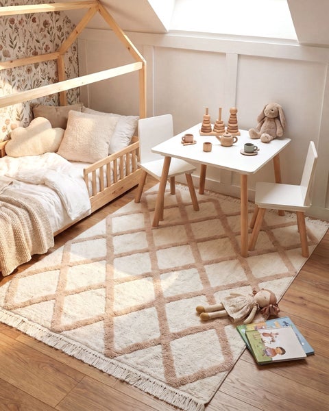 Ein gemütliches Kinderzimmer mit einem Hausbett, einem Kindertisch mit zwei Stühlen und einem Teppich mit Rautenmuster auf Holzboden.