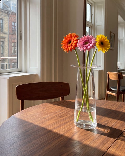 Drei bunte Gerbera in einer gläsernen Vase auf einem runden Holztisch. Der moderne Raum mit großen Fenstern wirkt hell und organisiert.