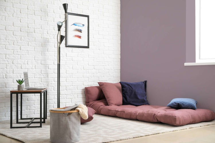 Modernes Wohnzimmer mit mauvefarbener Wand und weißer Ziegelwand. Bodenmatratze, Kissen, Stehlampe und Beistelltisch auf einem hellen Teppich.