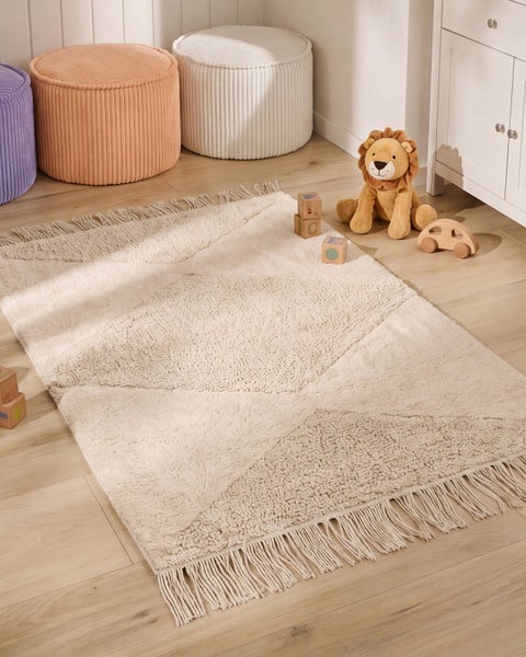 Ein rechteckiger Teppich mit Fransen und geometrischem Muster in einem hellen Kinderzimmer mit Spielzeug und Sitzhockern auf einem Holzboden.