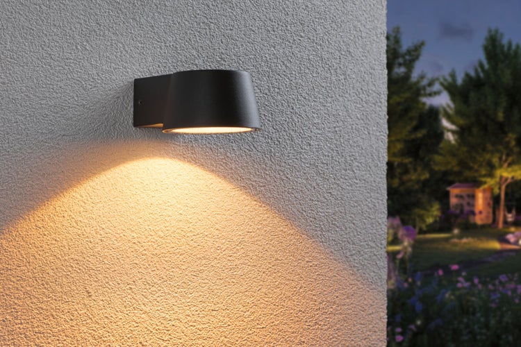 Moderne Außenwandleuchte an einer strukturierten Wand, strahlt warmes Licht nach unten ab, im Hintergrund ein Garten bei Dämmerung.