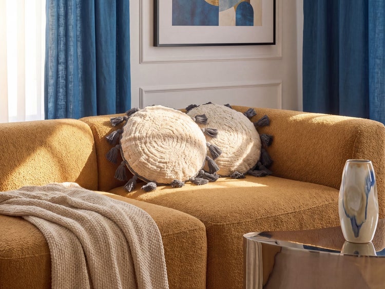 Modernes Wohnzimmer mit zwei runden Zierkissen mit Quasten auf einem senfgelben Sofa, einer Strickdecke und blauen Vorhängen.
