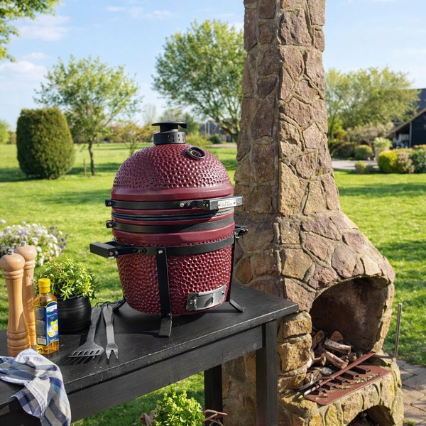 Roter Patton Kamado-Grill aus Keramik auf einem schwarzen Tisch in einem hellen Garten neben einem steinernen Außenkamin.