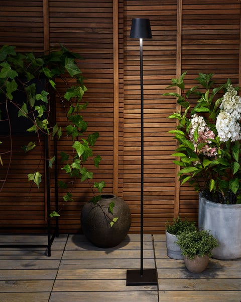 Moderne Außenstehleuchte in Schwarz mit schmalem Standrohr und konischem Schirm auf einer Holzterrasse zwischen verschiedenen grünen Pflanzen.