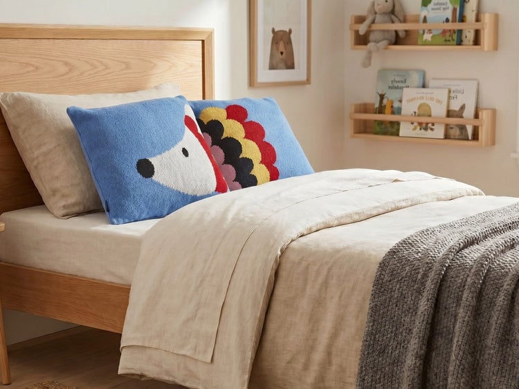 Helles Kinderzimmer mit einem Holzbett, heller Leinenbettwäsche und einem blauen Strickkissen mit buntem Igel-Motiv.