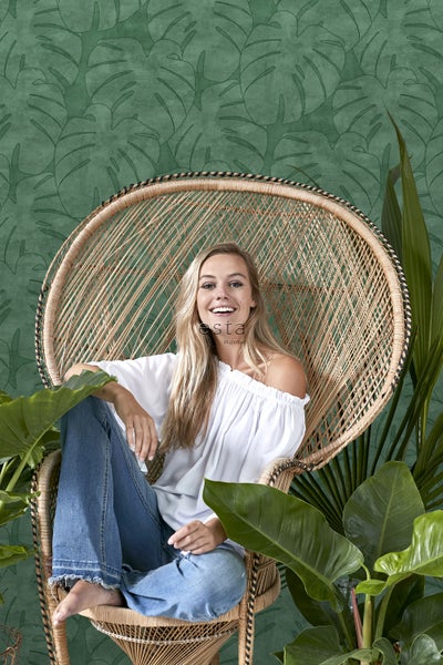 Frau sitzt in einem Rattansessel vor einer grünen Blättertapete.