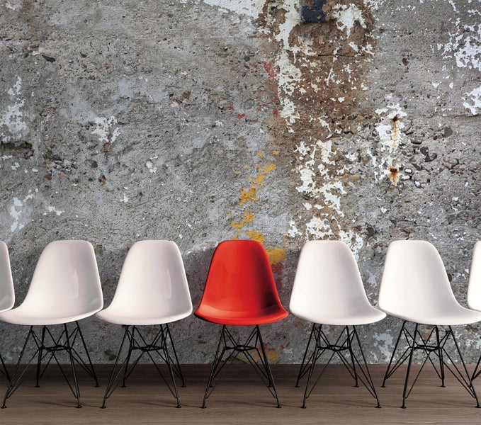 Fototapete mit Motiv einer verwitterten Betonwand und einer Reihe von Designerstühlen, darunter ein roter Stuhl als Akzent.