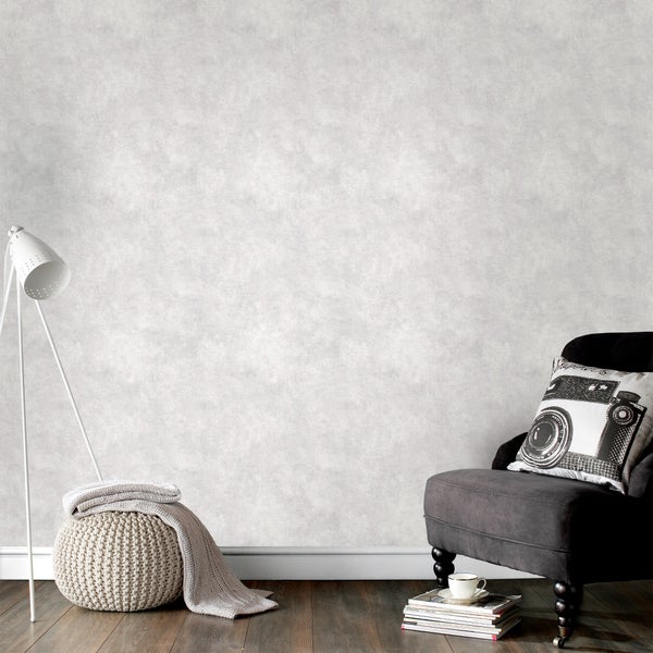 Wohnzimmer mit Vliestapete in Betonoptik, grauem Sessel, weißer Stehlampe und Strick-Pouf auf dunklem Holzboden.