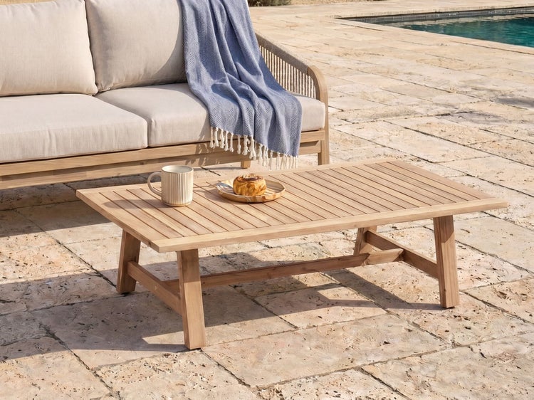 Rechteckiger Loungetisch aus Holz mit Lattenoberfläche auf einer hellen Steinterrasse neben einem Sofa und Pool.