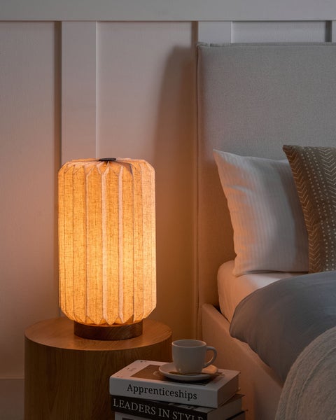 Zylindrische Tischleuchte mit plissiertem Textilschirm und Holzsockel auf einem runden Nachttisch aus Holz neben einem Bett.