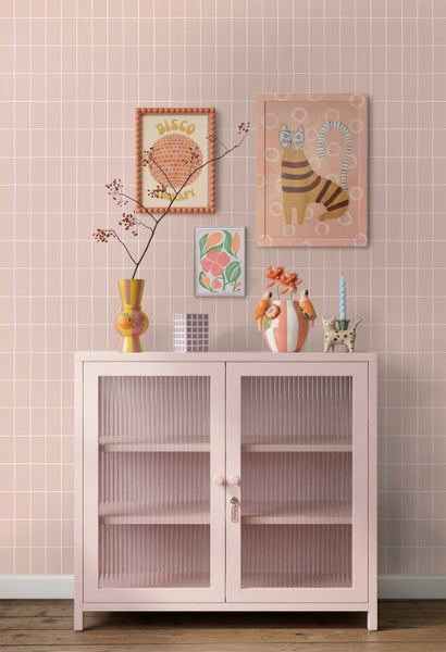 Rosa Metallschrank mit geriffelten Glastüren vor einer rosa gefliesten Wand, dekoriert mit bunten Vasen und Wandbildern.