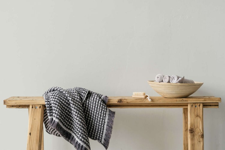 Eine rustikale Holzbank mit einem gemusterten grauen Handtuch, einer kleinen Holzbürste und einer Holzschale mit aufgerollten Handtüchern.