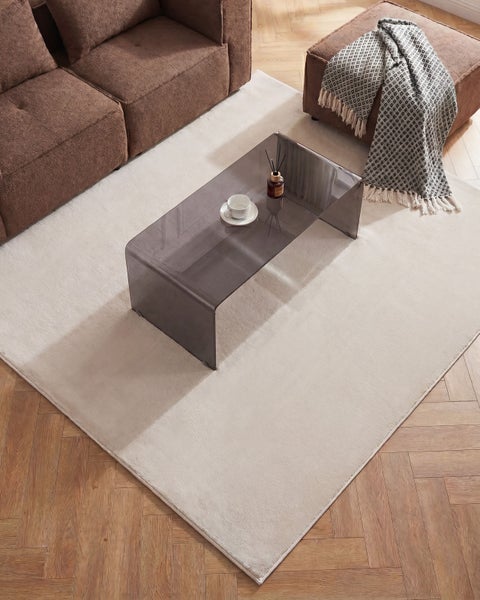 Rechteckiger, heller Teppich in einem Wohnzimmer auf Fischgrätparkett mit grauem Glastisch und braunem Sofa.