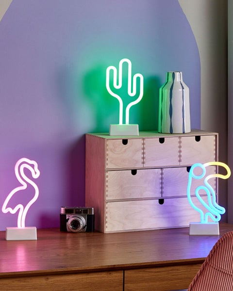 Heller Schreibtisch mit dekorativen LED-Leuchten im Neon-Stil in Flamingo-, Kaktus- und Vogelform, einem Holz-Organizer und einer Kamera.