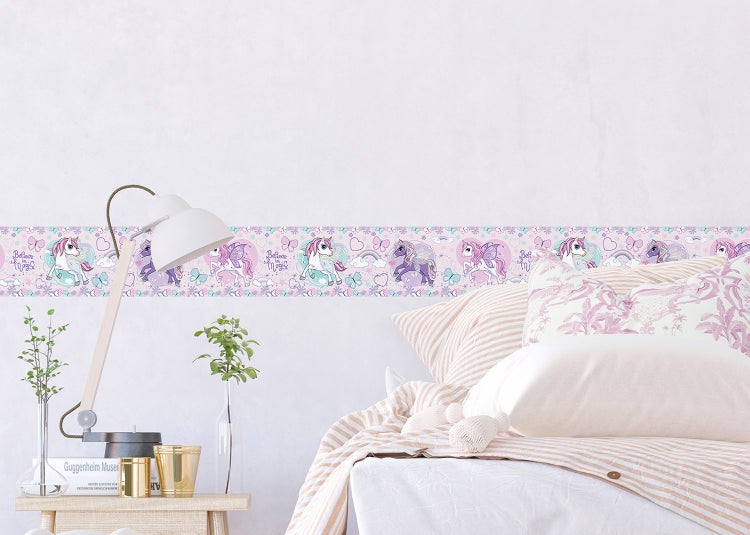 Helles Kinderzimmer mit Tapetenbordüre mit Einhornmotiven, Sternen und Schmetterlingen. Neben dem Bett ein Nachttisch mit Lampe und Pflanzen.