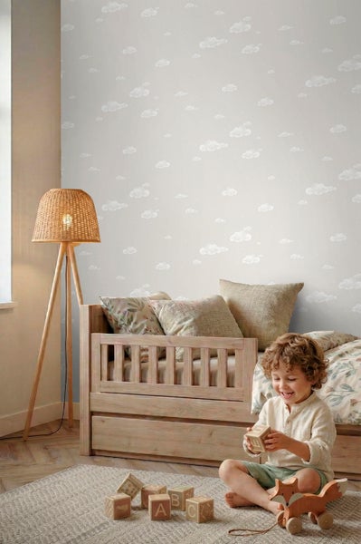 Tapete mit Wolkenmuster in einem hellen Kinderzimmer mit Holzbett, Stehlampe aus Geflecht und einem spielenden Kind.