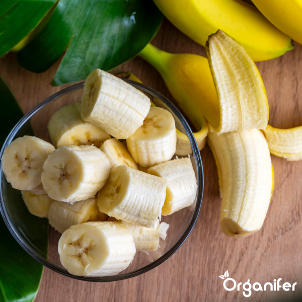 Geschnittene Bananen in einer Glasschale und eine geschälte Banane auf einer Holzoberfläche. Organifer Logo.