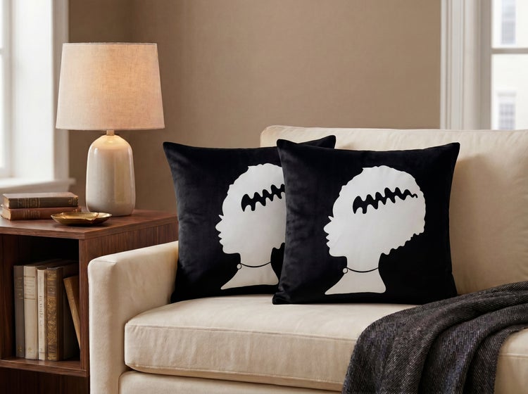 Zwei schwarze Zierkissen mit weißem Frauenprofil-Motiv auf einem hellen Sofa neben einem Beistelltisch mit Lampe.