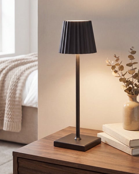 Moderne, schlanke Metall-Tischleuchte mit plissiertem Schirm auf einem Holztisch neben Büchern und einer Vase.