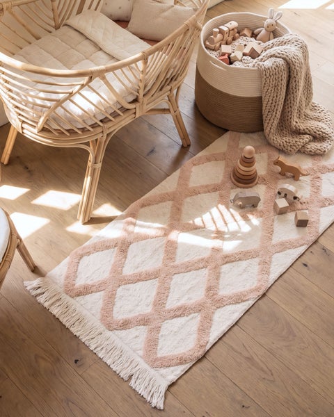 Ein Kinderzimmer mit einem Rattankinderbett, einem Aufbewahrungskorb, Holzspielzeug und einem gemusterten Läufer mit Fransen auf Holzboden.