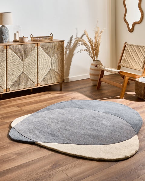 Moderner, organisch geformter Teppich in Grau und Creme auf Holzboden in einem hellen Zimmer mit Sideboard aus Holz und Flechtwerk.