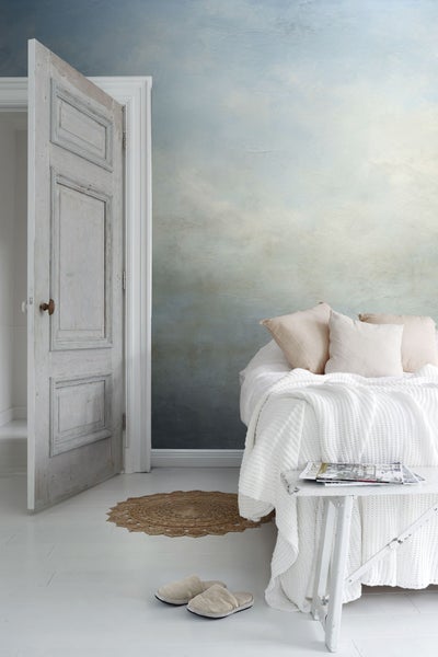 Helles Zimmer mit Bett, Beistelltisch, Teppich und geöffneter Holztür.