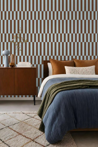 Ein Schlafzimmer mit einer Mustertapete mit vertikalen Streifen, einem braunen Sideboard, einem Bett mit Decken und Kissen sowie einem Teppich.