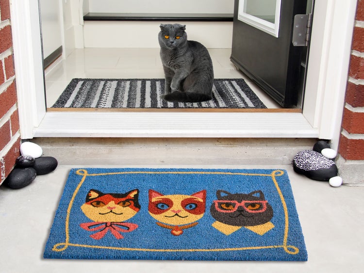 Fußmatte mit Katzenmotiven vor einer Haustür, mit einer grauen Katze im Hintergrund