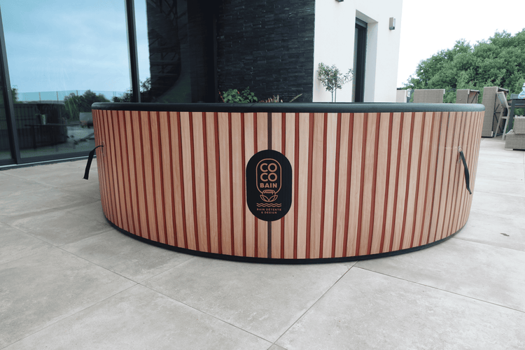 Runder Whirlpool mit vertikalen Lamellen in Holzoptik und Coco Bain Logo auf einer gefliesten Terrasse.