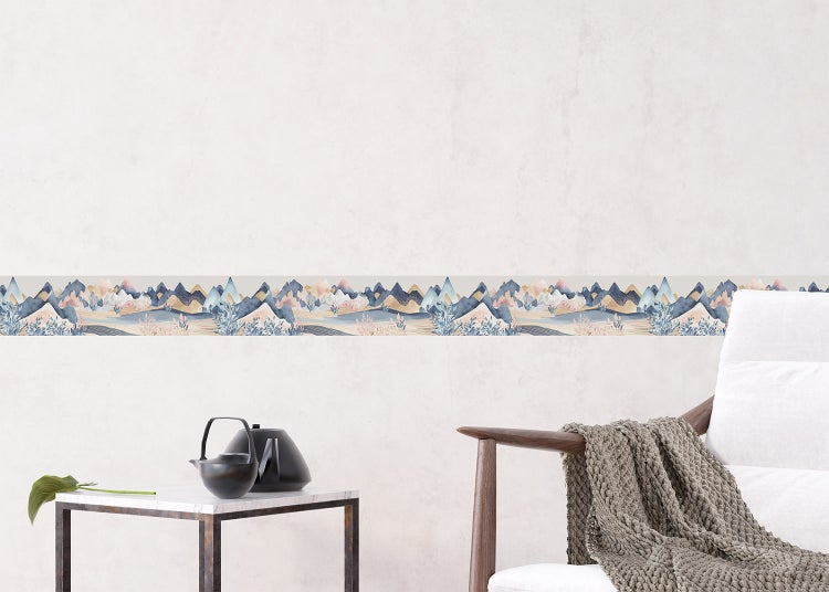 Selbstklebende Tapetenbordüre mit Berglandschaft in Aquarell-Optik an einer hellen Wand in einem Wohnzimmer mit Sessel und Beistelltisch.