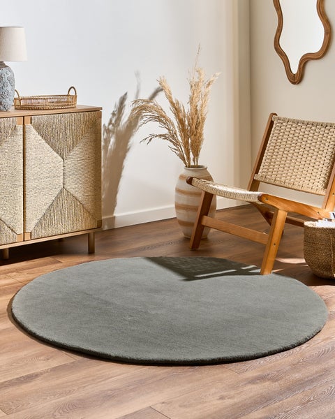 Hell gestalteter Wohnraum mit rundem grauen Teppich auf Holzboden, Holz-Sideboard mit strukturierten Fronten, geflochtenem Sessel und Bodenvase.