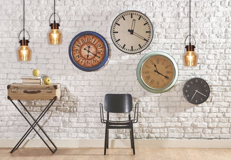 Dekoratives Wohnambiente mit Pendelleuchten, Vintage-Koffer auf Beistelltisch, Wanduhren und Stuhl vor einer Ziegelwand