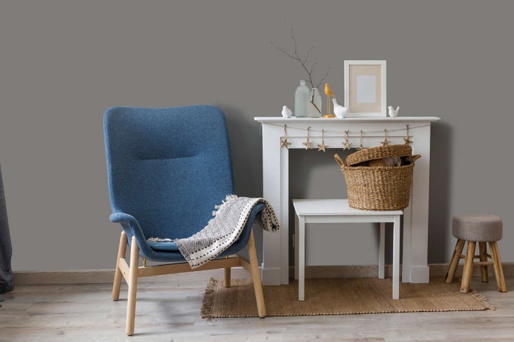 Modern eingerichtetes Wohnzimmer mit grau gestrichener Wand, blauem Sessel mit Holzbeinen, weißer Kaminkonsole und dekorativen Elementen.