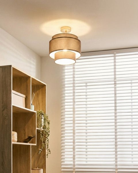 Moderne Deckenleuchte mit zweistufigem Textilschirm in einem hellen Wohnraum neben einem Holzregal und einem Fenster mit Jalousien.