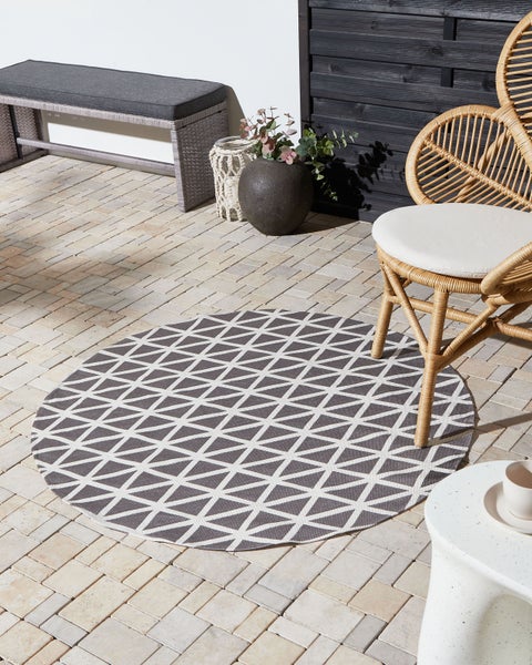 Runder Outdoor-Teppich mit grau-weißem Muster auf Steinterrasse mit Rattansessel, Bank und Beistelltisch. Helle und organisierte Außenanlage.