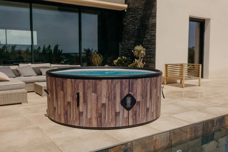 Runder Whirlpool in Holzoptik auf einer hellen, modernen Terrasse neben einem Pool mit Gartenmöbeln und großen Glasfronten im Hintergrund.