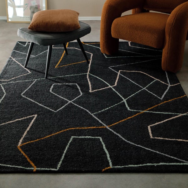 Moderner dunkler Teppich mit geometrischem Muster aus farbigen Linien in einem Wohnzimmer mit Hocker und Sessel.