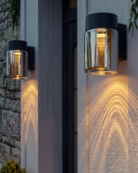 Zwei moderne Außenwandleuchten mit zylindrischem Glasschirm, die warmes Licht nach unten auf eine Hauswand werfen.