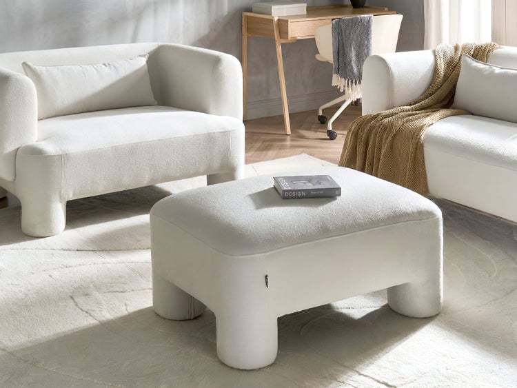 Moderner Polsterhocker aus weißem Bouclé-Stoff in einem hellen, organisierten Wohnzimmer mit Sofas, Schreibtisch und strukturiertem Teppich.