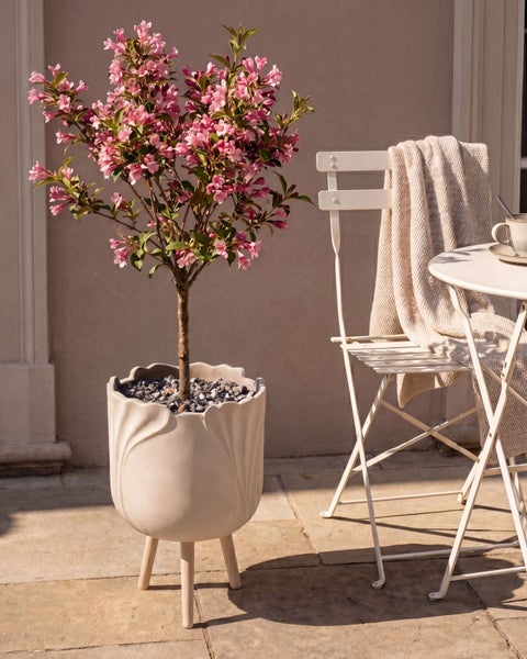 Blühende Weigela in weißem Dekotopf mit Holzfüßen auf sonniger Terrasse neben weißem Bistrostuhl mit Decke.