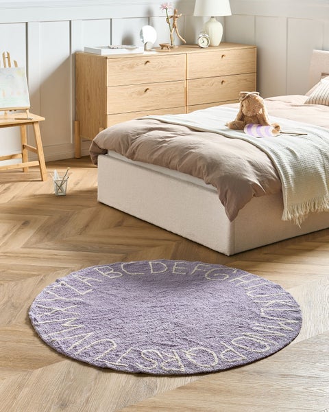 Runder Kinderteppich mit Alphabet-Muster in einem hellen Kinderzimmer mit Bett, Holzkommode und Parkettboden.