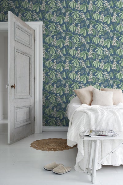 Schlafzimmer mit einem Kachelofen, einem Bett mit einem geflochtenen Kopfteil, einem Nachttisch und einer Wandtapete mit Blattmuster